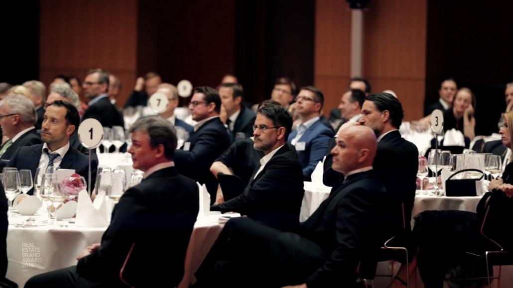 You can see a seated audience in a large hall listening to a speaker who is also shown in the event movie.