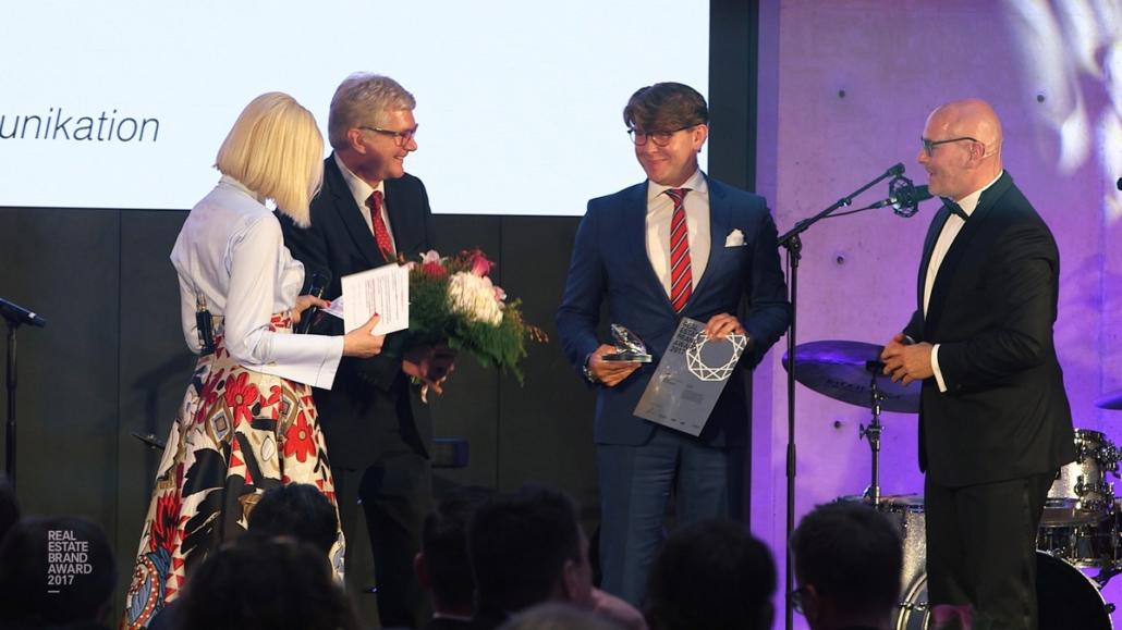 This picture shows in a medium long shot the award ceremony of an event participant, who holds a certificate and the prize in the form of a diamond in his hands.