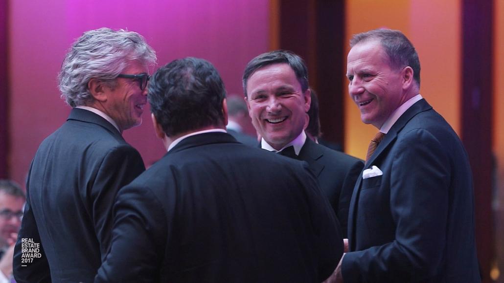 In a medium close-up shot, we see four entrepreneurs in black suits against an out-of-focus background, talking excitedly.