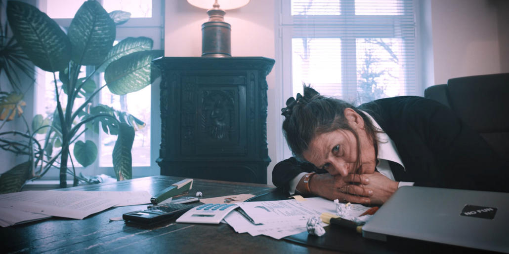 We see the actress Gundi-Anna Schick in a semi-close-up as she is bent over her large, wooden desk with a dissolved look, resting her head on her clasped hands. She looks anxiously past the camera on the left. The room is flooded with daylight. This is a still of a movie production.