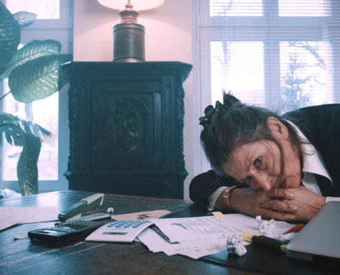 We see the actress Gundi-Anna Schick in a semi-close-up as she is bent over her large, wooden desk with a dissolved look, resting her head on her clasped hands. She looks anxiously past the camera on the left. The room is flooded with daylight. This is a still of a movie production.