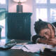 We see the actress Gundi-Anna Schick in a semi-close-up as she is bent over her large, wooden desk with a dissolved look, resting her head on her clasped hands. She looks anxiously past the camera on the left. The room is flooded with daylight. This is a still of a movie production.