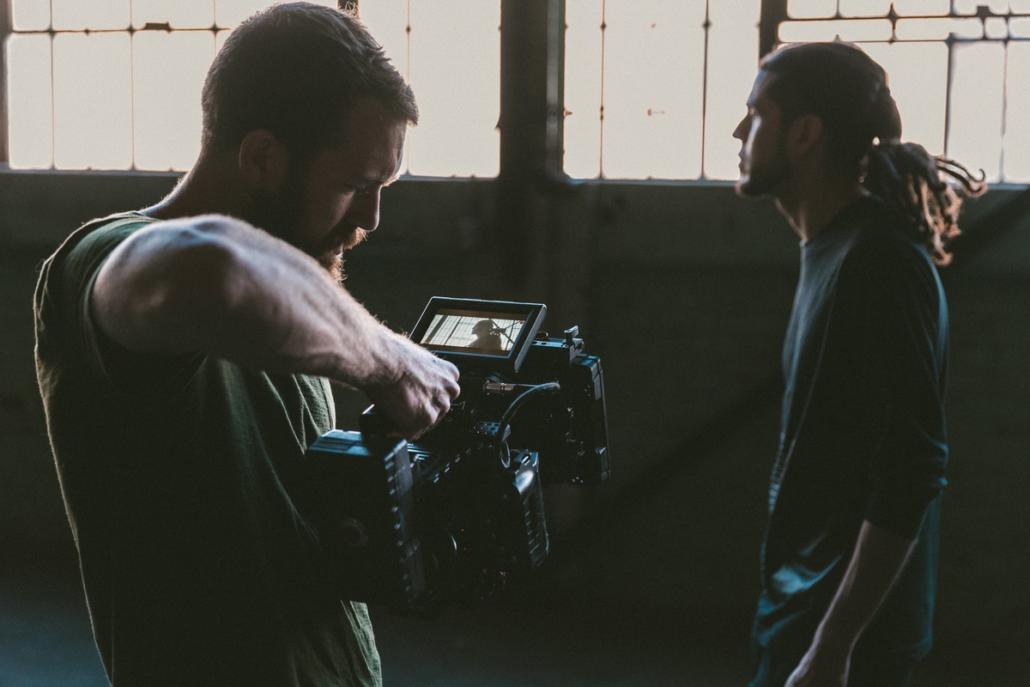 Camera operator of the film production company berlin is shooting a middle aged protagonist in front of a factory wall with a RED camera from a lower angle and there are windows in the background