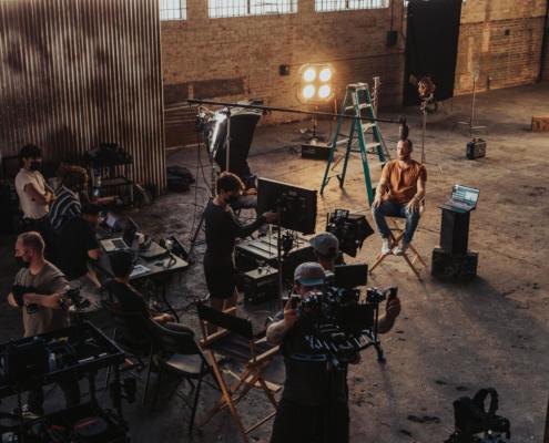 Viele Mitarbeiter vom Filmunternehmen Berlin in der Drehpause am Set eines Interviewdrehs, von schräg oben fotografiert und in einem alten leeren Raum eines Backsteingebäudes