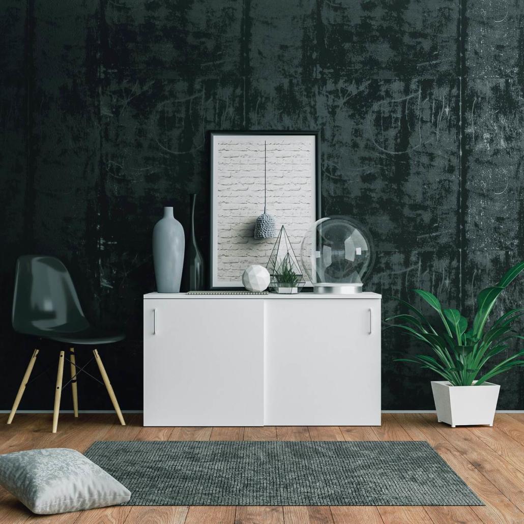 Frontally we see a 3D render of a modern interior. A rustic, black stone wall in the background in front of which a white chest of drawers stands in the center of the image. To the left of it stands a modern chair, to the right of it a green plant in a white pot. On the dresser are glass shapes and a vase in mouse gray color. Directly above it is a black framed picture. The picture shows a black mysterious writing on white paper.