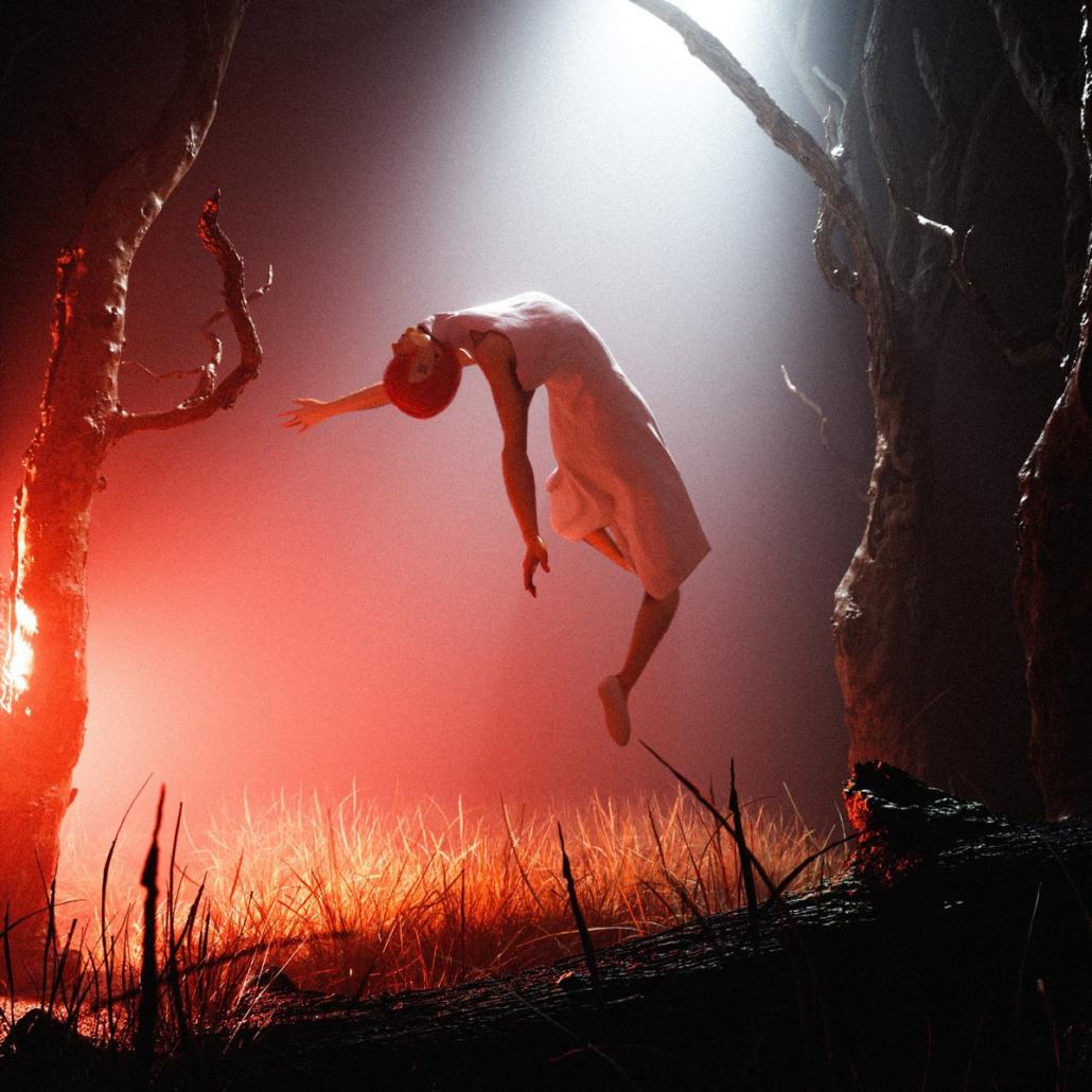A 3D render shows a lifeless woman floating towards the sky in front of a dark background between leafless trees as part of the work of a 3D animation studio of Berlin. The ground consists of grass and in the foreground a tree trunk protrudes horizontally into the image. A strong red light shines on the woman from the left as well as a white light coming from the sky in the form of a cone of light.