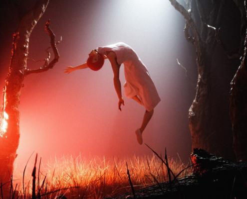 A 3D render shows a lifeless woman floating towards the sky in front of a dark background between leafless trees as part of the work of a 3D animation studio of Berlin. The ground consists of grass and in the foreground a tree trunk protrudes horizontally into the image. A strong red light shines on the woman from the left as well as a white light coming from the sky in the form of a cone of light.