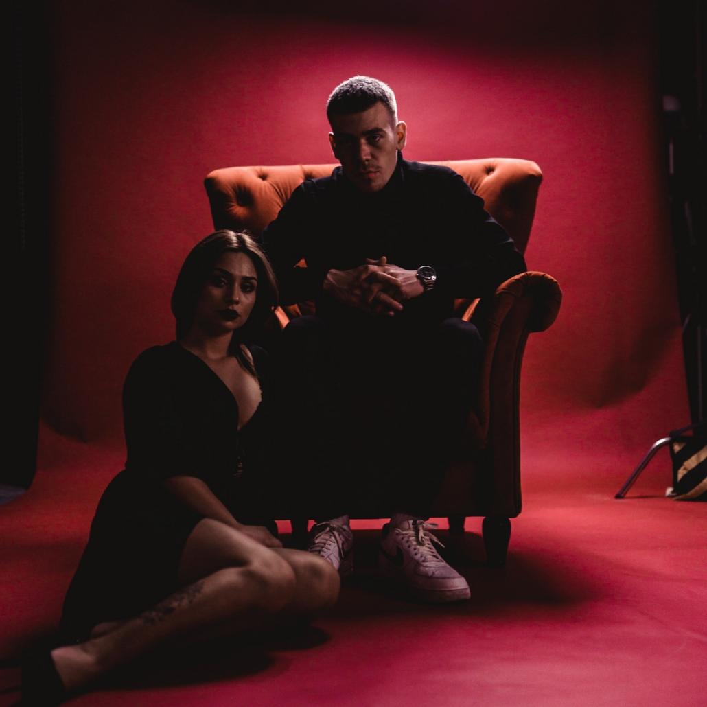 If you want to create a music video, this type of illustration shown here can be useful: On this picture, against a red background, you can see an actor sitting in the middle of an orange armchair. He has short black hair and is wearing a black turtleneck sweater. He has his hands clasped and wears a noble wrist watch around his left arm. Slightly tired, he looks past the camera at the top left. Directly to his right, we see a young woman with long black hair sitting on the floor with bent knees and bold, dark red lipstick. Her emotionless gaze resembles that of the man.