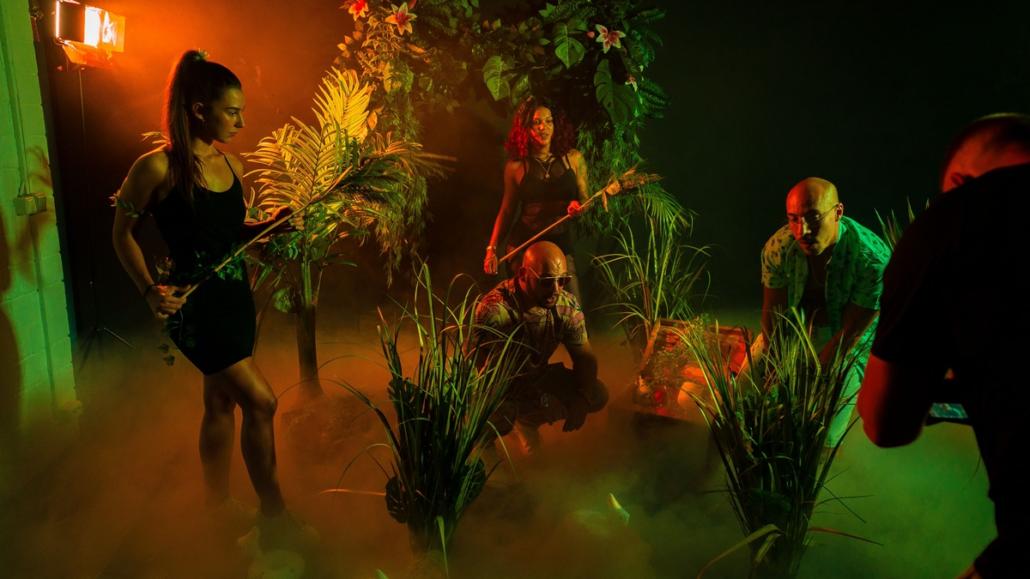 A group of artists, consisting of two men and two women, can be seen. A man kneels in the center of the picture in a Hawaiian shirt, with a bald head and sunglasses. Behind him and to his left is a young woman in a black dress with a plant stem in her hands, held like a guitar. On the right, slightly crouching, we see a bald man with glasses and mustache, also dressed in a Hawaiian shirt. This is a good way to create a music video, using impressive green-orange effect lighting and a fog machine, as shown here. In the front right of the picture, we see the cameraman cropped in as he holds his camera and is recording.