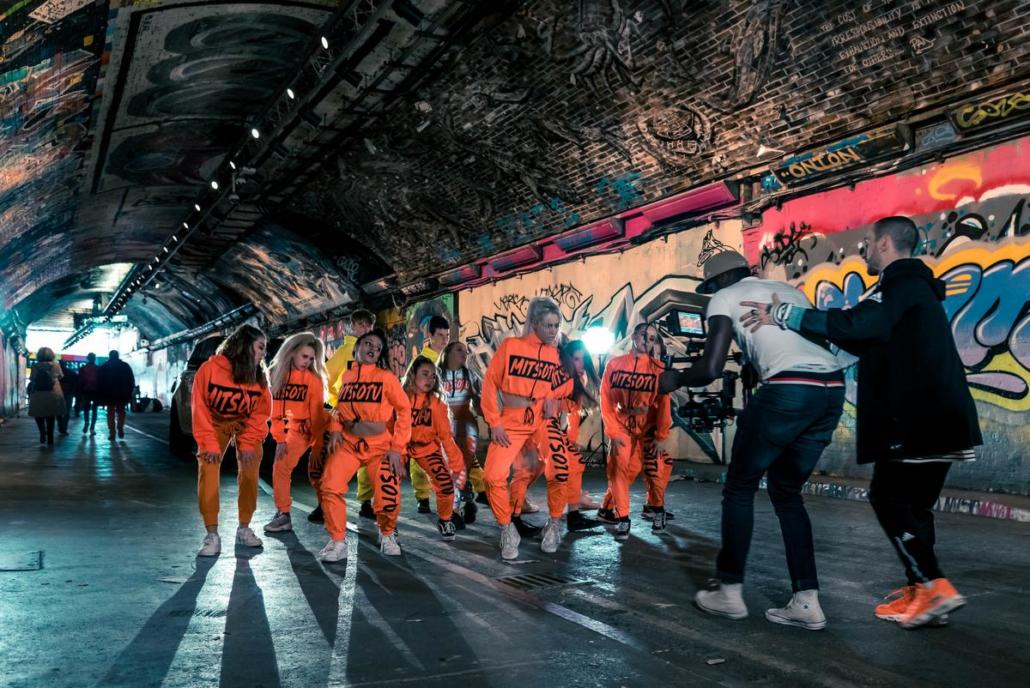 It is interesting to create a music video shot in a graffiti-strewn tunnel, as shown in this image: we see a group of young girls in orange clothing in a dead shot. Together they perform dance choreography in front of the camera. The cameraman can be seen on the right of the picture with a camera gimbal and is being held and guided by another man for safety reasons during his shot.
