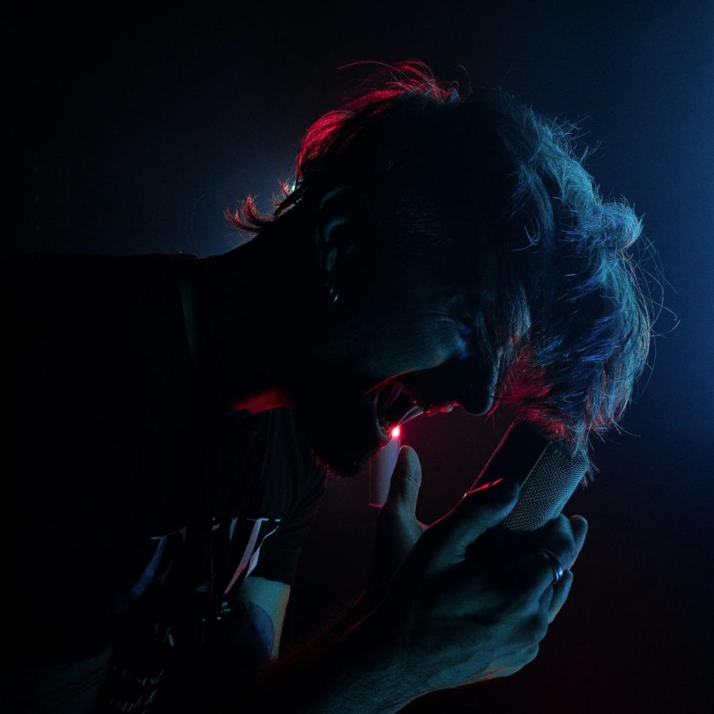 In the picture we see the face of a front singer bent forward, centered in the picture and in profile. Against a dark background we see him singing powerfully into his microphone. From the background he is illuminated by a red neon tube. If you want to create a music video, this image is a good inspiration.