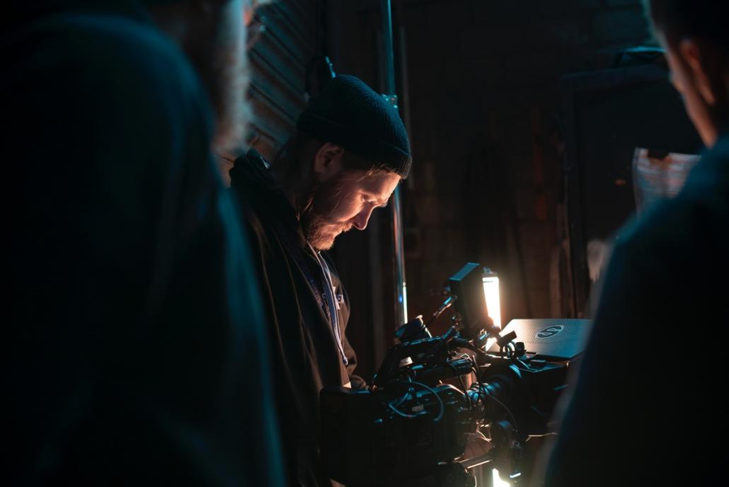 This picture shows the cameraman at a film production in Berlin in the semi-close-up, who is looking through the control monitor of his film camera in order to evaluate the recorded image. On the left and right in the front of the picture, we see two other crew members in the blur.