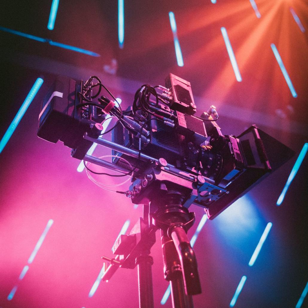 This illustration impressively shows, from a frog's-eye view, a ready-to-go, elaborately rigged film camera, which is equipped with a large V-mount battery. The camera is on a sturdy tripod in a film studio, whose ceiling is equipped with several turquoise neon lights and the room shines in volumetric light.