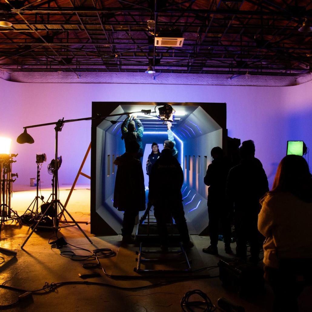 In a studio, fictional film formats can be realized very well, as illustrated in this photo: We see the crew of a film production in Berlin, which is just preparing the appropriate light for the next take. The light on the set is magenta and bluish. In the middle of the set is a fictional, futuristic backdrop in front of which the actor is standing, shot by a camera that is on a dolly.