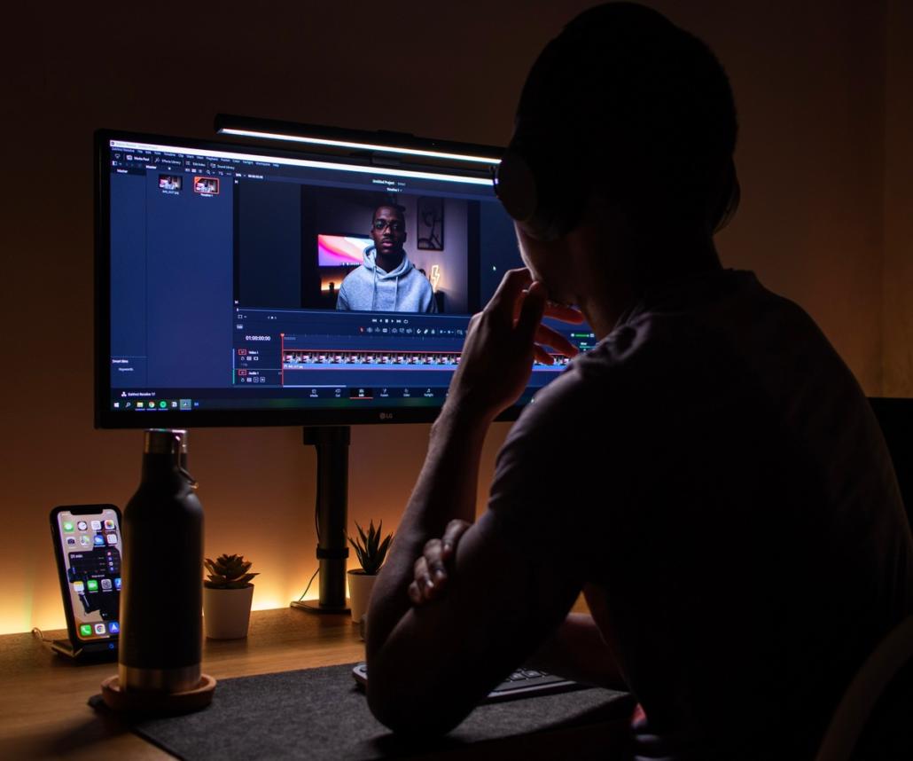 We see a film editor sitting in front of his digital editing program, in a slightly oblique perspective from behind. He is examining a picture and has assumed a pensive pose with his left hand and propped up elbow. The background of the editing suite is slightly illuminated by a warm artificial light and the rest of the room is kept rather dark.