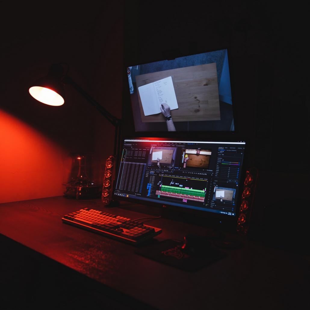 We see a very stylish and neatly set up editing suite, diagonally from the right. The focus of the view here is on a widescreen monitor that shows a well laid-out project structure of the editing program Adobe Premiere. Directly above the editing monitor is a large control monitor that shows the current program image. An ordinary desk lamp protrudes over the table on the left and bathes the image in a red hue.
