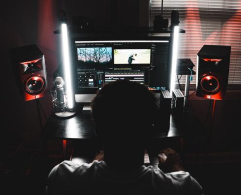 A stylized workplace of a film editor is shown on this image: We see the film editor directly from behind and slightly from above, sitting and working at his editing station. His workstation is very neatly arranged and is slightly illuminated by a red light from the floor. There is a widescreen monitor on the table, as well as two white glowing neon tubes arranged vertically on either side of the monitor and shining in the direction of the viewer. To the left and right of the table are speakers on stands.