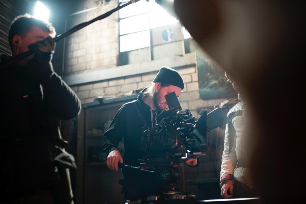 Create an image film with real film elements, as exemplified in this image: In the center of the image is a cameraman who is looking through the viewfinder of his camera while filming is in progress. To his left is the sound man with a boom in his hand, and to his right, cropped into the image, we see the director, who is following the recorded image through a control monitor.