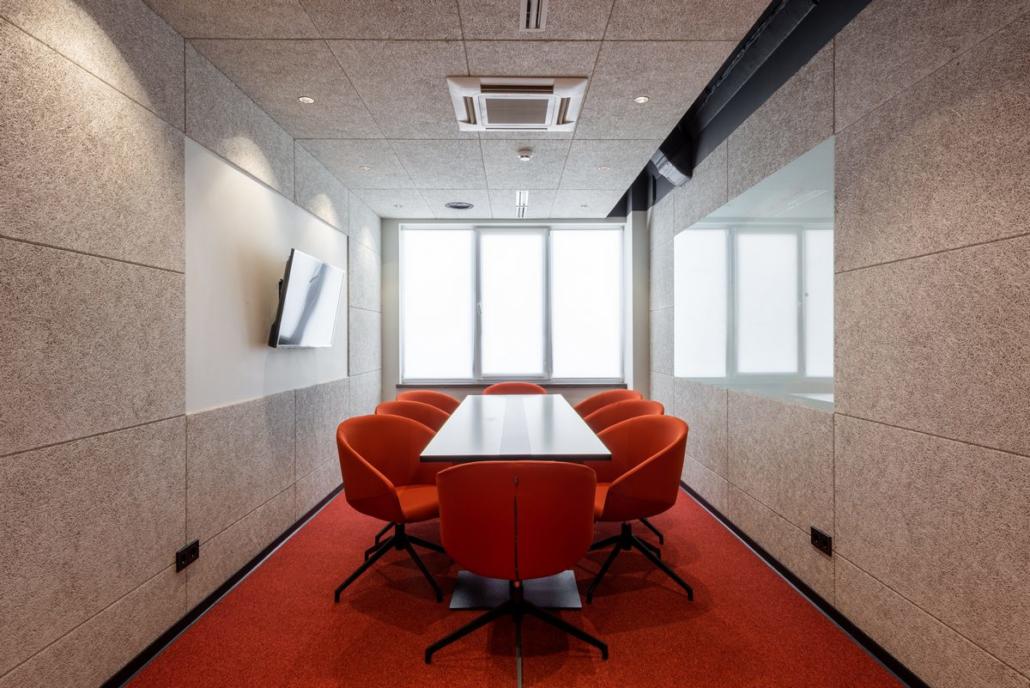 An Image film can show a meeting room as seen in this picture. We see a rectangular, modern meeting room with beige soundproof walls, a large flat screen on the left side and large windows at the back of the rear wall. The room is covered with red carpet and in the center is a large, long, rectangular table, which is surrounded by red chairs in a round design. The picture shows the room with table and chairs in perfect symmetrical arrangement.