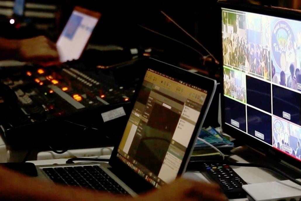 This picture shows the control room of a live stream company. We see a large program screen on the right, an open laptop with running streaming software in the center, a large mixing console in the blur further to the left, and another open laptop in the very back left of the image together with the hand of an employee. We also see the hand of another employee in the left foreground.