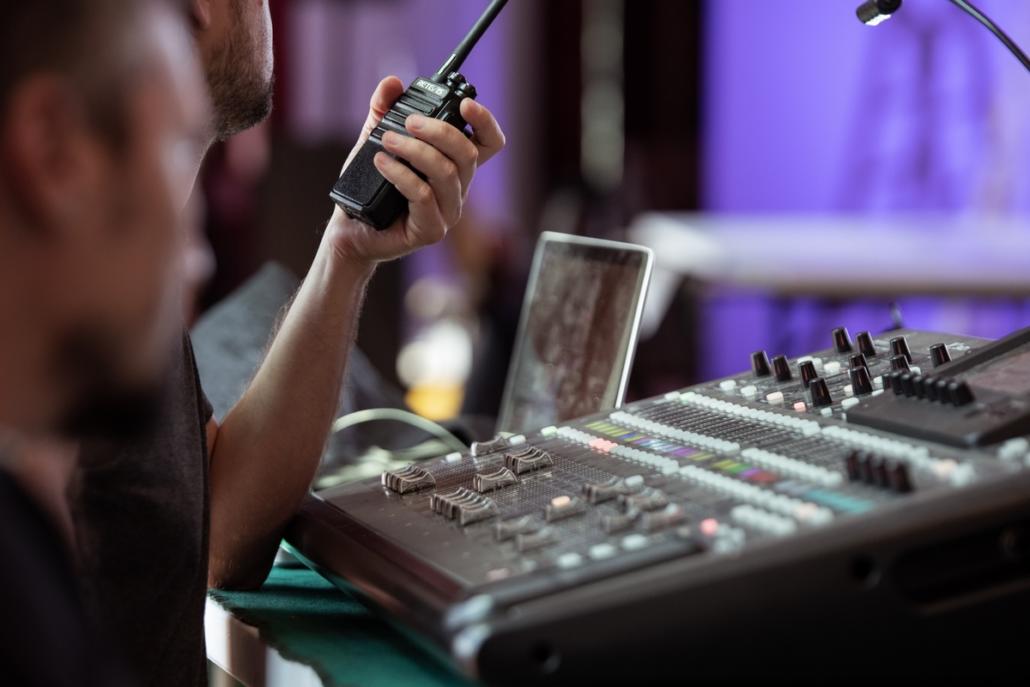 In the focus of the image we see a sound mixer and behind it a laptop in the blur. On the left in the crop and filmed in close-up from the side, we see two crew members of a live production. One employee is also in focus and is holding a walkie-talkie in his left hand. The other is seen out of focus over the shoulder in the front left.