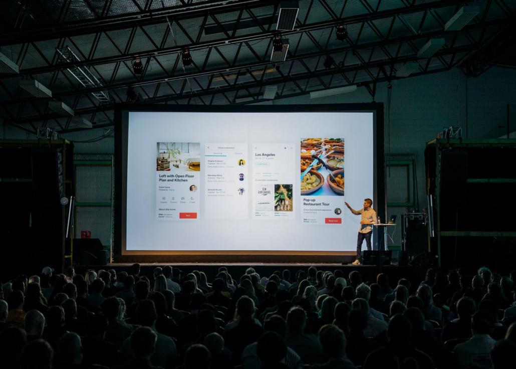 With a professional team from Berlin, a live production can be skillfully driven, as illustrated in this picture: We see a stage in the long shot from the front, on which a speaker is standing at a lectern on the right and is currently giving a PowerPoint presentation that can be seen on a large screen. In the foreground we see the audience from behind. The room is dark and only the stage is illuminated.