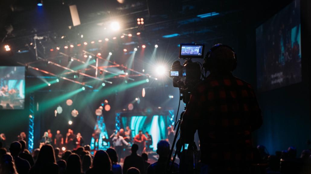 In the background, a large stage can be seen in the blur, which is equipped with many green and red stage spotlights that illuminate both the stage and the audience. The audience is in the middle ground of the image. Right in front we see, in sharp focus, in the foreground from behind the cameraman of a live stream company at his studio camera, which is on a tripod.