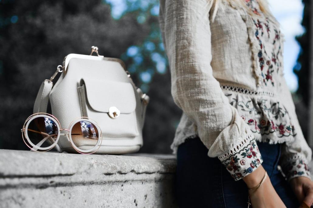 Product video, as shown in this shot, in which a branded ladies' handbag is placed on a stone wall, which is the focus of the viewer's attention. On the right side of the image we see a woman leaning against the wall and putting her glasses on the wall in front of the ladies handbag.