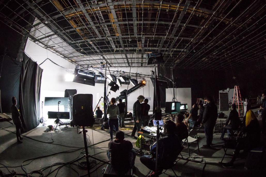 Shown is a very large film studio in a strong wide angle shot, diagonally left from the side. The set is illuminated with a lot of spotlights and equipped with a camera crane. The film set is surrounded by numerous sitting and standing personnel, who seem to be preparing for the shoot.