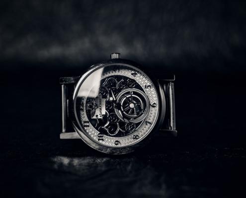 In close-up, we see the filigree, metal dial of a luxurious clock in the center of the image against a black background. The clock lies on a black, strongly reflecting background, in which a kind of mist is reflected, which can also be seen in the background. The scene is in black and white and the glass of the clock reflects light in an impressive way. This is an impressive way to have a TV spot created.