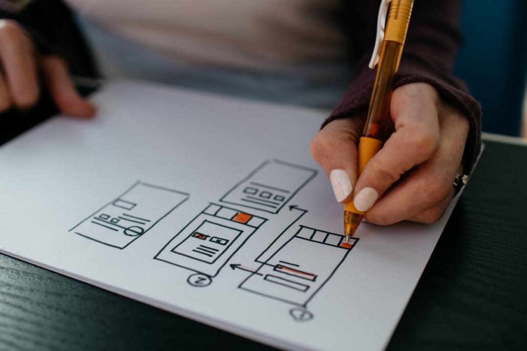 A close-up of a woman's left hand holding a ballpoint pen and sketching visualizations and processes on a white sheet of paper. If you want to produce an animated video explainer, preparatory conceptual work is very important.