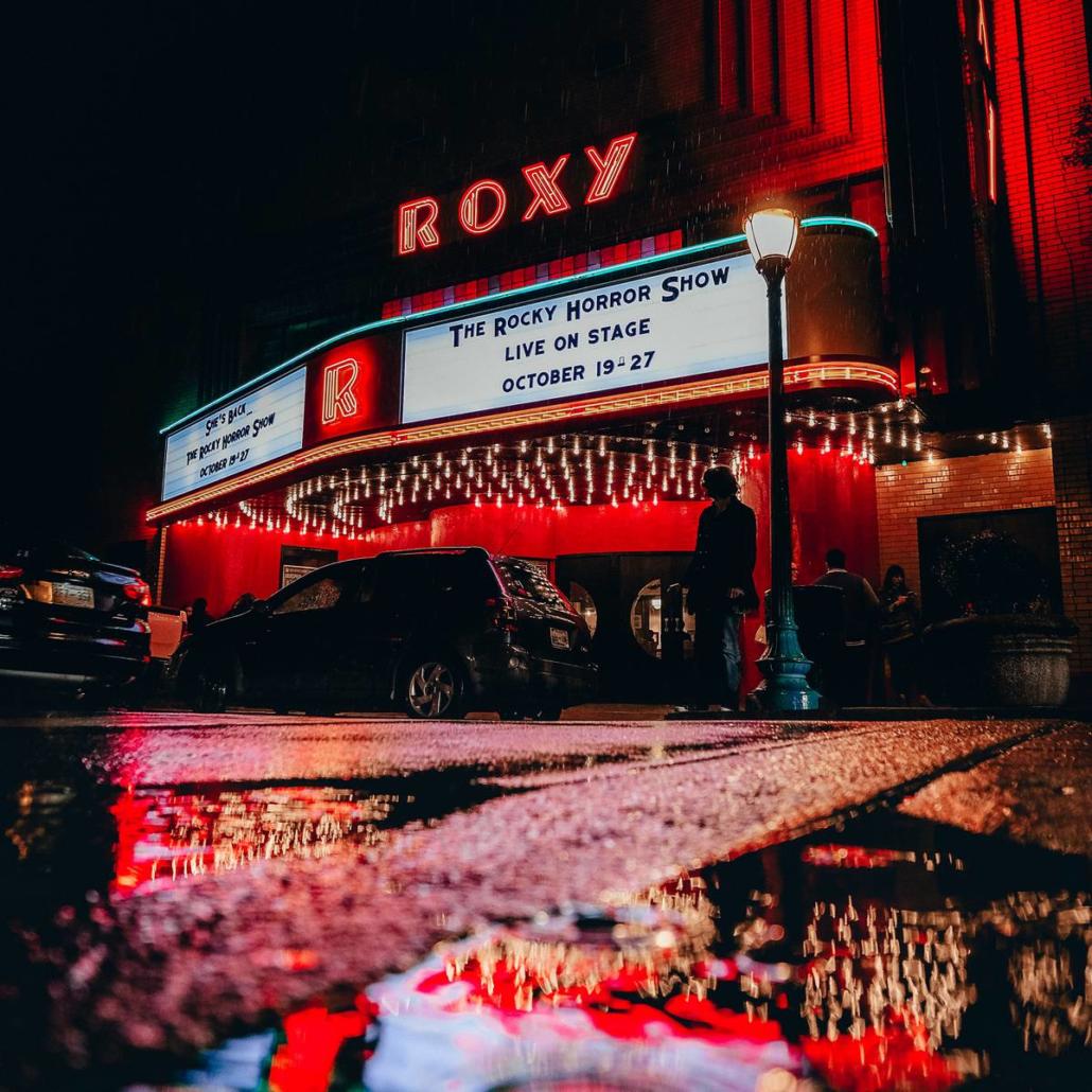 If you wanted to enjoy the 10 best movies of 2022 in a particularly breathtaking atmosphere, this cinema depicted here with its magnificently illuminated exterior façade would be an ideal place to do so: In frog perspective, we look from a wet street at the cinema, which is equipped with numerous lights. On the roof of the cinema, the word 