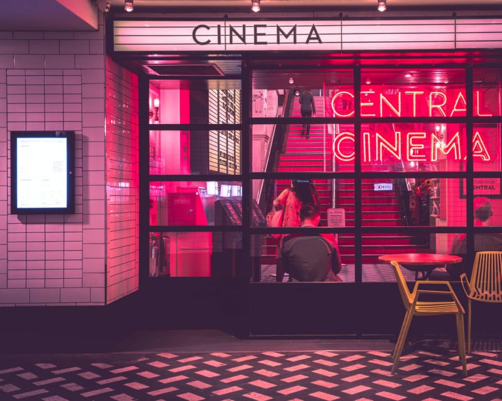 In the picture, the entrance of a red-lit cinema can be seen in a cutaway as a place where the 10 best movies of 2022 can be consumed excellently. The cinema is equipped with a large glass facade and a typical white advertising sign that reads 