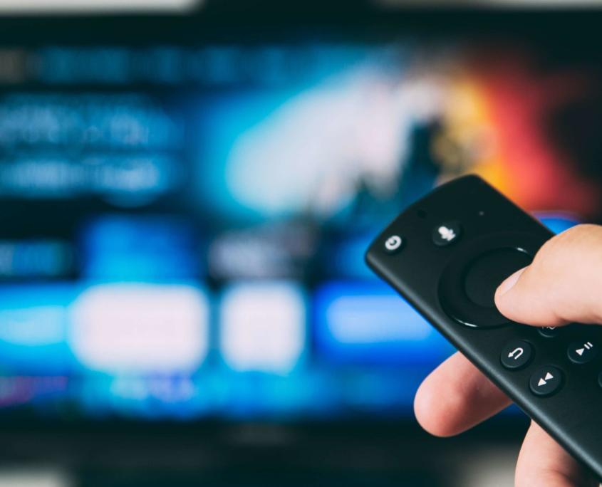 The 10 best series can be watched very well on a smart TV, as illustrated in this picture: We see a smart TV in the blur, which suggests the user interface of streaming service providers. In the foreground, we see a person's hand holding a remote control in focus on the right side.