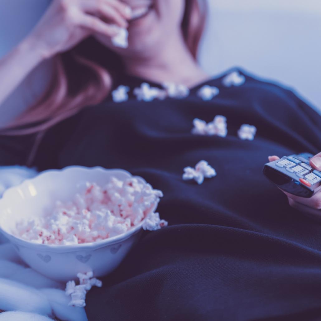 The picture shows a young woman in a blue top, sitting comfortably on the sofa, happily eating popcorn from a bowl with her right hand, while holding the remote control with her left hand and watching the best TV shows 2022. Her face is only cropped in the picture, so that only the lower half of her face is shown.