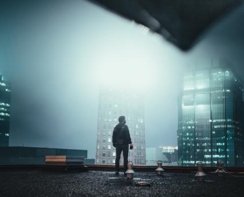 Shots of skyscrapers are a favorite in the best tv shows all time, as illustrated in this image: we see a man on a skyscraper roof from the bottom view. The sky is very foggy and the man is looking at opposite, even higher rising, glass skyscrapers, from whose direction a strong blinding, white, diffuse light is coming.