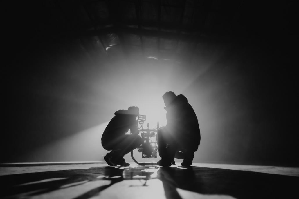 Two cameramen can be seen kneeling from behind and in black and white, who are currently in a video production in Berlin and together operate a camera gimbal.