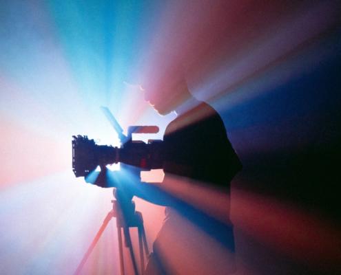 For a video production in Berlin, interesting light setups can be useful, as shown here: We see a cameraman standing at his camera and at the same time in an imposing backlight of red and blue color.