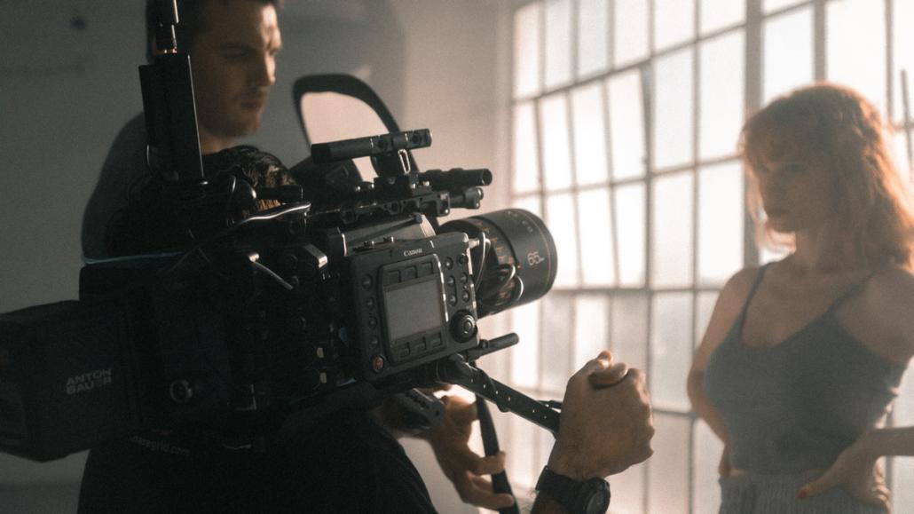 Hosting a video production in Berlin in a hall with high windows, as shown in this picture. Diagonally from behind we see a cameraman holding a rigged camera in his hands and filming an actress. To his left, the director is checking the image he has just recorded.