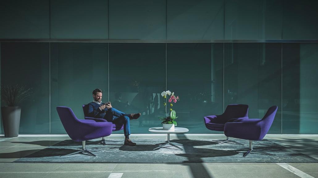 The image shows a long shot of a man in a blue sweater and jeans sitting elegantly in one of four purple armchairs arranged symmetrically around a round white table. Behind him is a large glass wall and behind it a turquoise background.