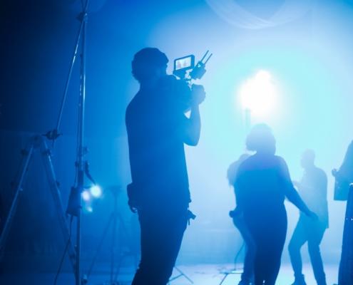 The picture centrally shows the cameraman of a video production company standing on a blue-lit film set with dancing actors in the background.