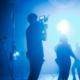 The picture centrally shows the cameraman of a video production company standing on a blue-lit film set with dancing actors in the background.