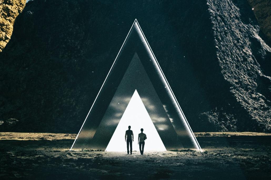 In the long shot, we see two people in a fictional environment walking toward a triangular structure that glows white.