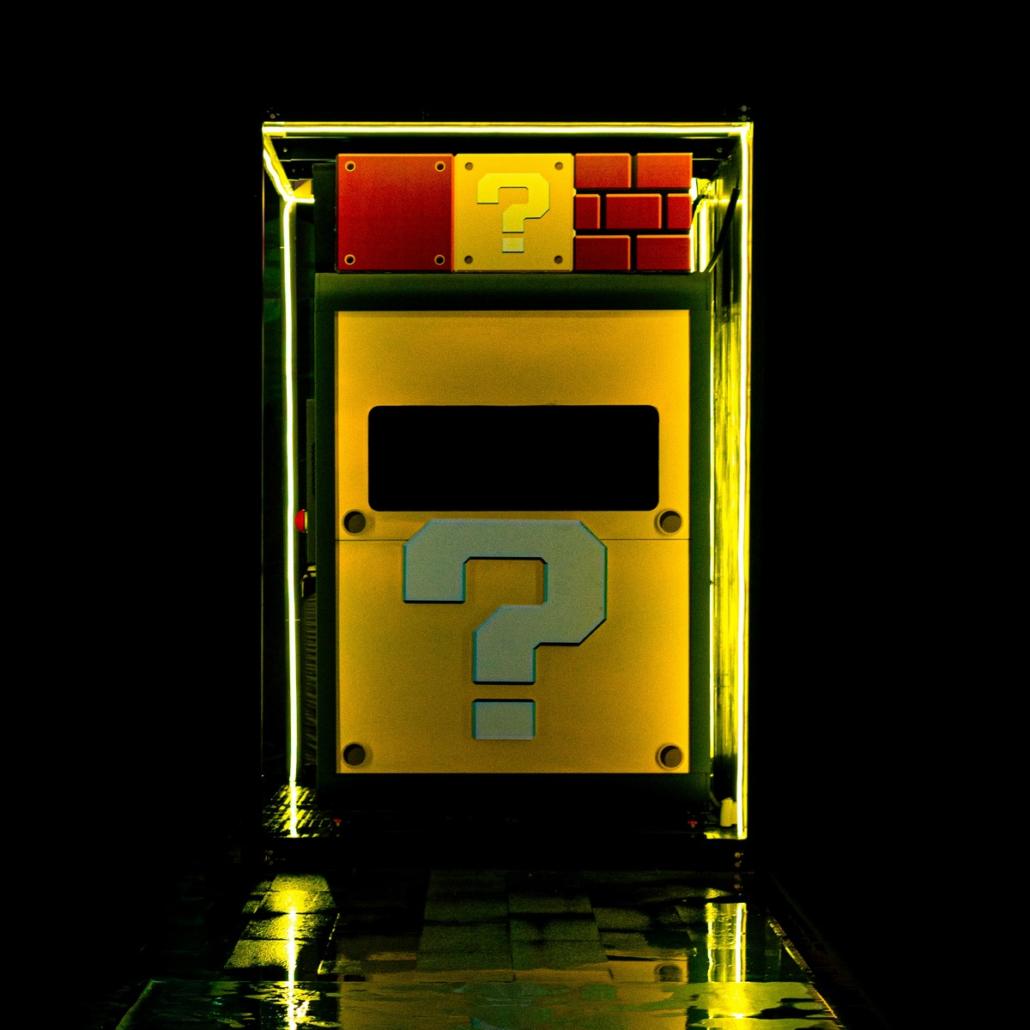 Against a black background we look at a structure consisting of yellow and red blocks, to which two question marks are attached. Around the square structure is an additional transparent block, which emits yellow neon light that is reflected on the floor.