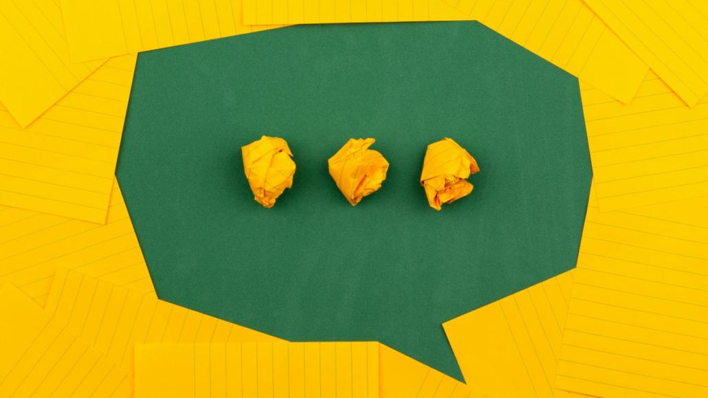 A speech bubble made of green paper can be seen in this image, which is the result of yellow index cards being pushed together. In the speech bubble you can see three folded index cards, which, in the form of three dots lying next to each other, symbolize the spoken text of an explainer video company.