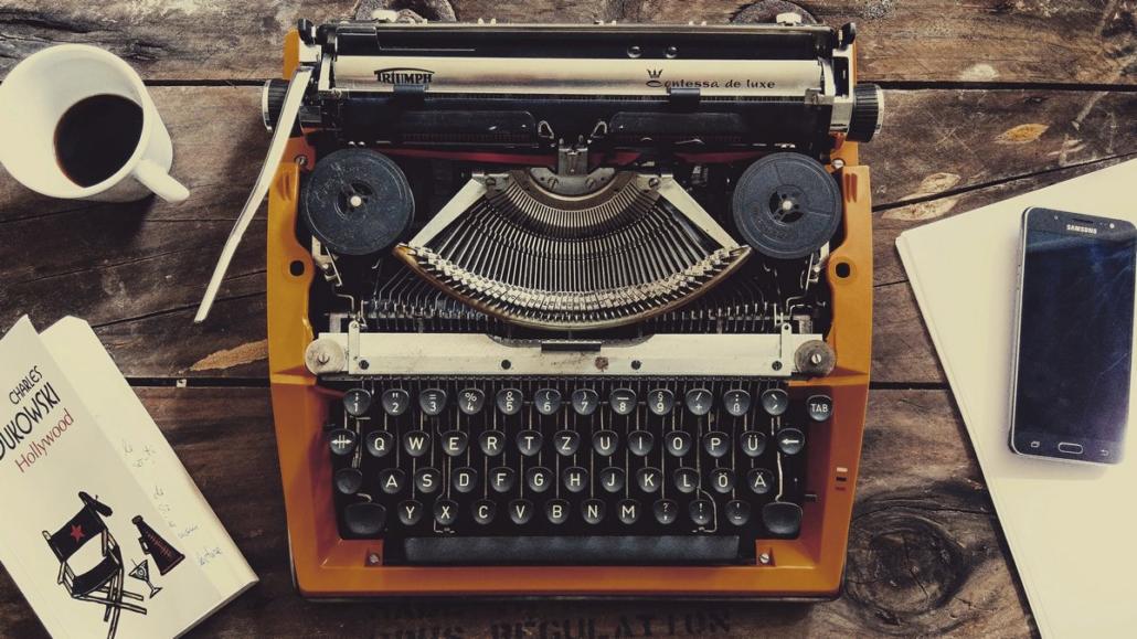 The top view shows a venerable typewriter standing on a wooden table. To the left of it lies a book. Above the book is a half-drunk cup of coffee. To the right of the typewriter lies a writing pad and on top of it a smartphone. This illustration is meant to illustrate the conception phase of an explainer video company.