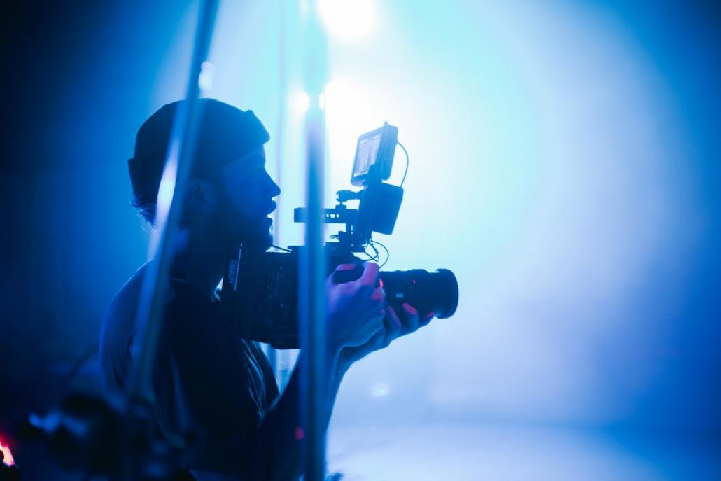 Pictured in this image is a cameraman in the semi-close-up, who is on a blue-lit set shooting a music video on behalf of a video agency.