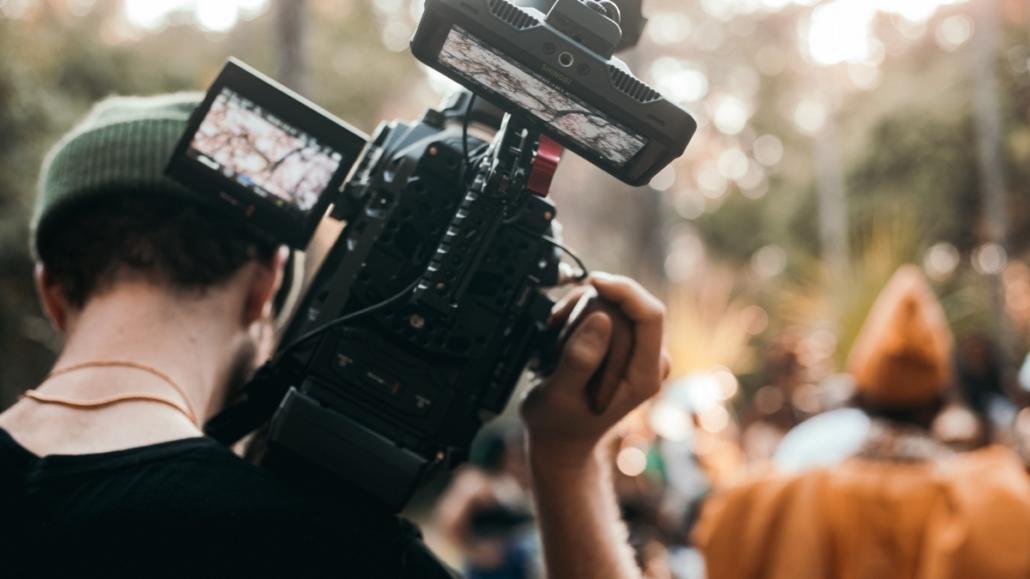 In this picture, the cameraman of a video agency can be seen from behind, carrying his camera on his shoulder. In the background we see several people out of focus in the open air.