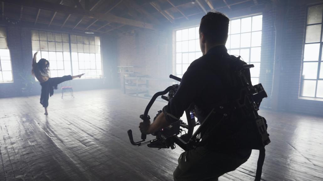 In a large light-flooded room, a dancer can be seen at the back on the left side, who is performing a dance with her leg stretched out and is being filmed by a cameraman from a video agency, who is at the front right of the picture.